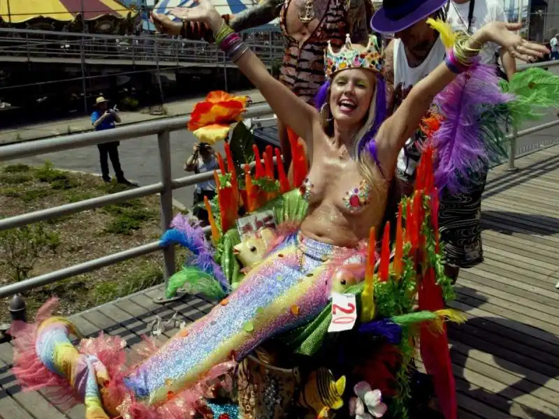 MERMAID HISTORY a rainbow mermaid wearing a crown is pushed down the boardwalk in some sort of crustacean chariot in 